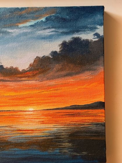 A close up angle of my orange sunset painting, highlighting the texture of the canvas and the rich, layered colors of the clouds and their reflection on the water.