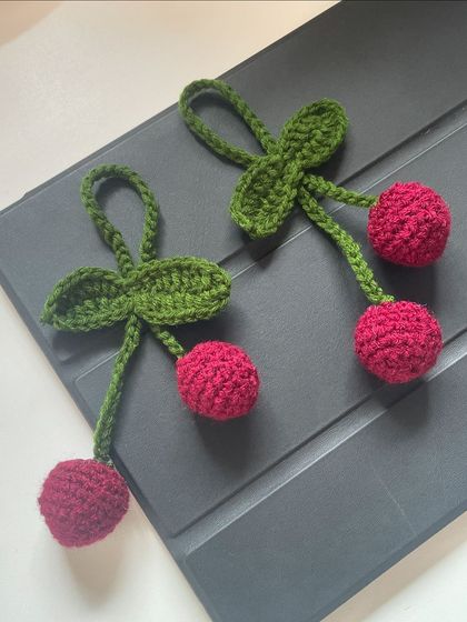 A pair of sweet cherry bag charms. Hand-crocheted with bright red yarn, these are perfect for adding a touch of retro fun to your accessories.