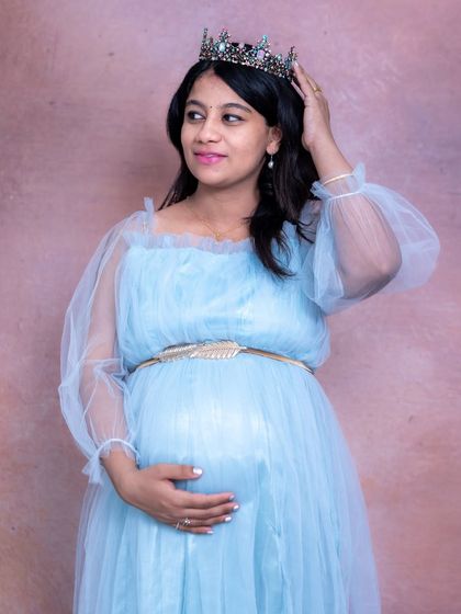 A regal portrait of a mom-to-be adjusting her crown, wearing a beautiful light blue gown in a studio setting.