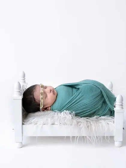 A newborn swaddled in a teal wrap, sleeping soundly in a miniature white bed against a clean, high-key background.