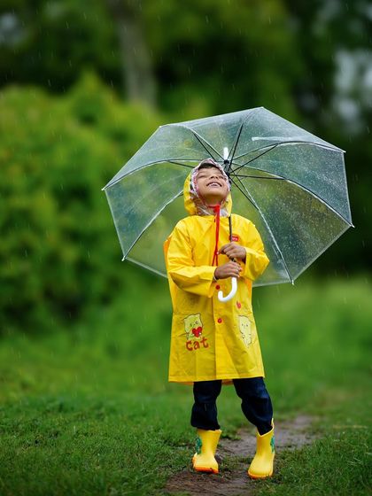The pure joy of playing in the rain! A little boy in a yellow raincoat and boots looks up at the sky with a happy smile.