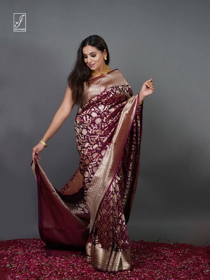 A studio shot for a brand's 'Riwaayat Collection,' featuring a model in a deep magenta Banarasi silk saree. The pose is designed to display the full drape and rich golden zari work of the fabric.