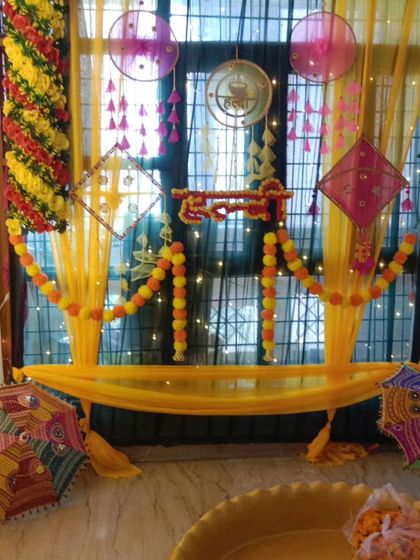 A window space transformed into a beautiful Haldi decoration corner. The setup uses yellow drapes, marigold garlands, and Rajasthani umbrellas to create a festive photo spot.