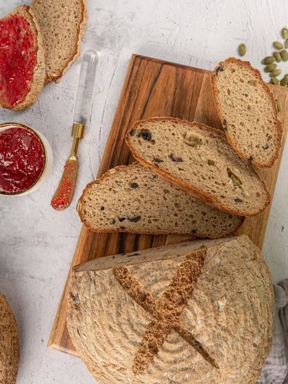 A rustic loaf of sourdough bread, sliced to show the beautiful crumb structure. The styling with jam and a wooden board gives it a wholesome, artisanal feel.