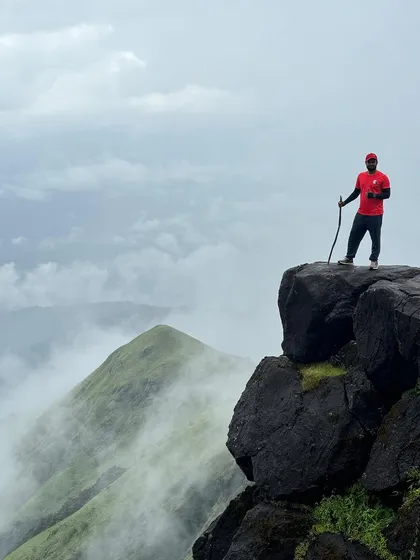 A trekker stands on the edge of a cliff on the Kumara Parvatha trail, with the misty Shesha Parvatha in the background. The views on this trek are simply surreal.