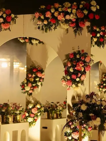 A beautiful reception entrance featuring a wall of modern white arches, each adorned with cascading arrangements of pink and red roses, creating a romantic and fragrant welcome.
