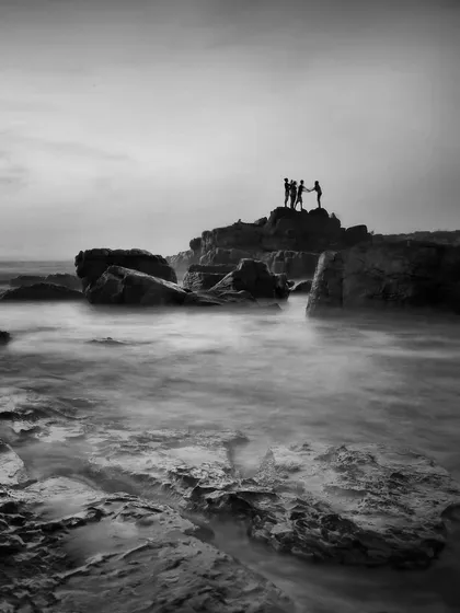 Silhouetted figures stand atop a rocky outcrop as the sea swirls around them. The long exposure blurs the water, contrasting its softness with the hard edges of the rocks.