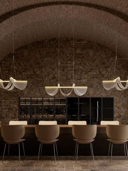 Three Drape Lights by Jamie Wolfond for Moooi are installed over a long dining table in a rustic, brick-arched hall. The gentle curves of the draped material soften the industrial feel of the space, providing a warm and elegant illumination.