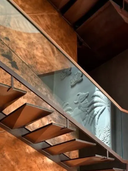 A close-up of the sculptural staircase, showing the cantilevered metal treads and the reflection of the carved wall panel in the glass balustrade.