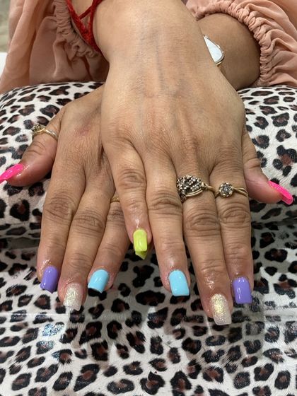 A playful and vibrant multi-color manicure. Each nail is painted with a different pastel shade, including neon yellow, lavender, and sky blue, with glitter accents for a fun and eye-catching result.