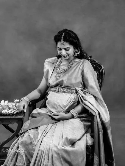 A timeless black and white portrait of an expecting mother in a silk saree. She is seated gracefully, adorned with traditional jewelry, looking down at her baby bump.