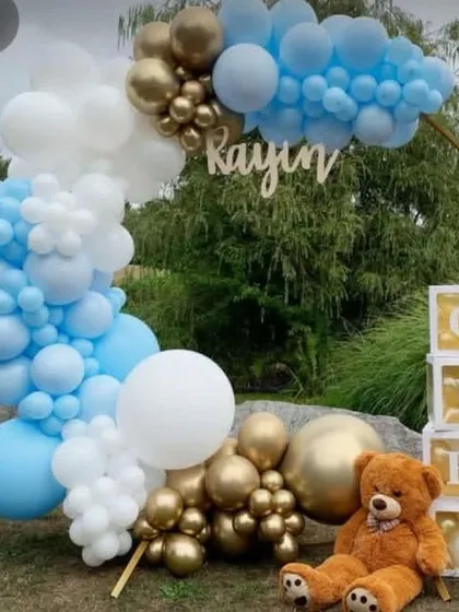 An outdoor teddy bear theme for a first birthday. This setup includes a balloon arch in blue, white, and gold, 'ONE' blocks, and a cute teddy bear.