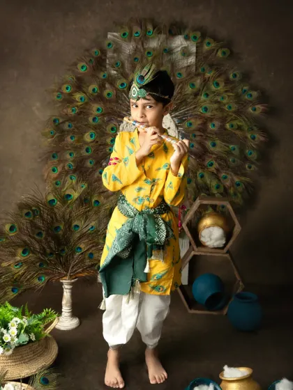 A boy dressed as Lord Krishna stands proudly playing a flute, with a backdrop of peacock feathers creating a divine atmosphere.