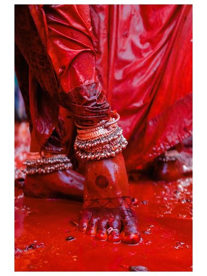 A close-up detail shot from Holi, focusing on feet adorned with traditional anklets, standing in a pool of red-colored water. It's an abstract but powerful image of the festival's essence.