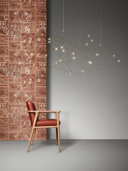 The linear version of the Flock of Light is suspended next to an armchair, its form resembling a glowing branch or a constellation.