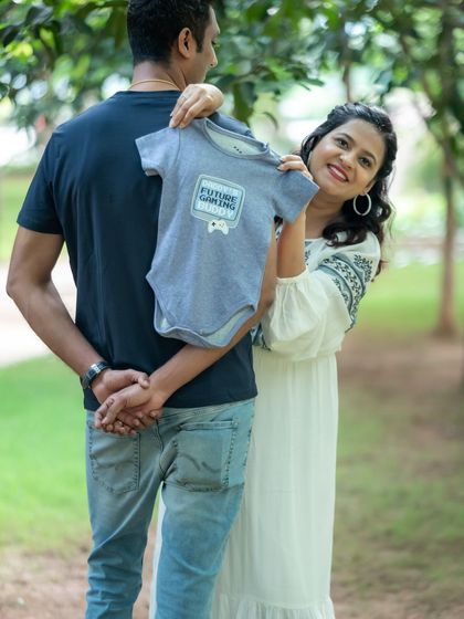 A playful and sweet announcement photo. The mom-to-be holds up a "Future Gamer" onesie behind her partner's back, a cute surprise captured during their outdoor session.