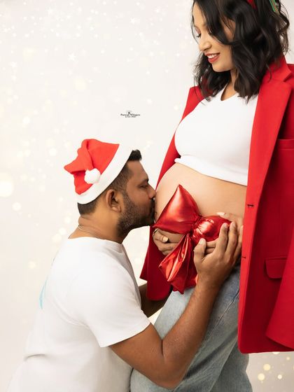 A heartwarming and creative Christmas shot. The husband-to-be kisses his wife's belly, which is adorned with a big red bow like the most precious gift.