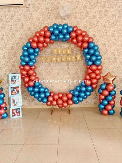 A balloon ring decoration for a baby's rice ceremony (Annaprasana). The setup uses red and blue balloons with 'BABY' blocks and star foil balloons.