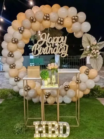 An elegant outdoor birthday decoration with a balloon ring in neutral tones. The setup is completed with display stands and a light-up "HBD" sign, perfect for a garden or backyard party.