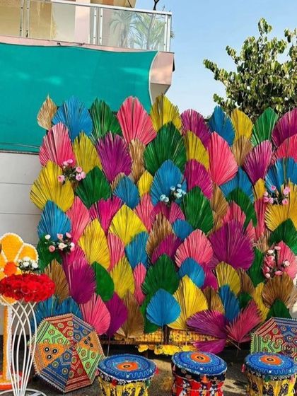 A modern and artistic photo booth backdrop made of colorful, overlapping palm leaves. The setup is completed with printed stools and quirky props for fun pictures.