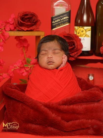 A fun and quirky "Black Label" themed newborn shoot. This is for parents who want something unique and humorous. The baby is wrapped in red, surrounded by props.