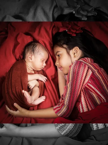 A powerful and artistic shot of an older sister lying beside her newborn sibling, captured in a selective color style to emphasize the deep red tones and the emotional connection.