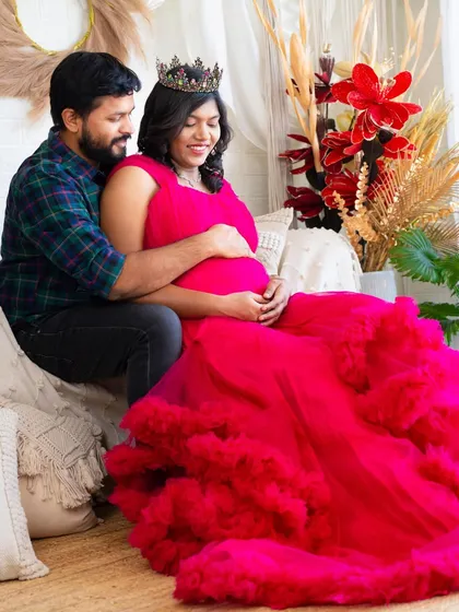 A tender moment on our studio couch. The partner embraces the mom-to-be from behind as she wears a vibrant pink gown, both sharing a quiet, happy moment.