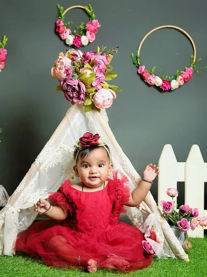 A sweet baby girl in a red dress sits in front of a lace teepee decorated with flowers. This boho-chic setup is both elegant and playful.