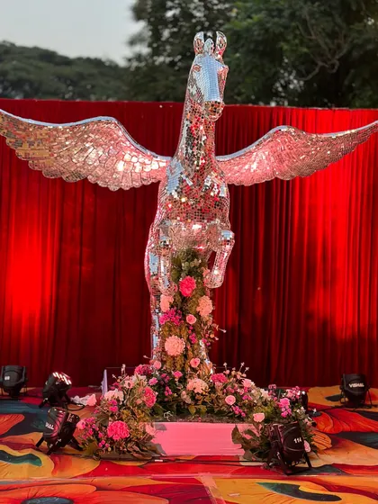 A custom-made, life-sized Pegasus statue covered entirely in mirror pieces, serving as a spectacular centerpiece for a glamorous event. The statue is surrounded by pink flowers, catching the light and creating a dazzling focal point.