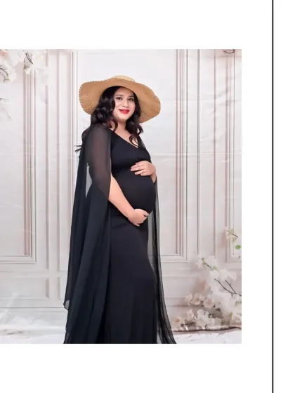 A stylish solo portrait featuring a chic black bodycon dress with a sheer cape and a fashionable straw hat. The simple, textured white backdrop keeps the focus on the modern and elegant silhouette.