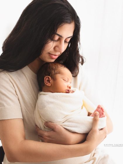 A mother holds her sleeping newborn, swaddled in white. The soft window light and her gentle gaze create a serene and emotional portrait.