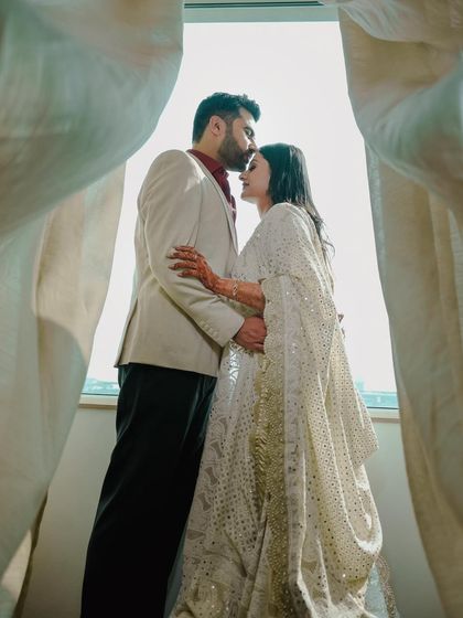 A beautifully framed silhouette of a couple by a window, capturing a private moment as he kisses her forehead, with soft light filtering through the curtains.