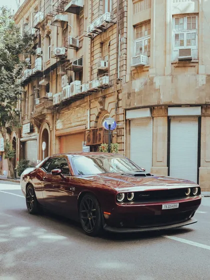 A modern muscle car, the Dodge Challenger, looks right at home on the historic streets of Bombay, its deep red color standing out against the stone buildings.