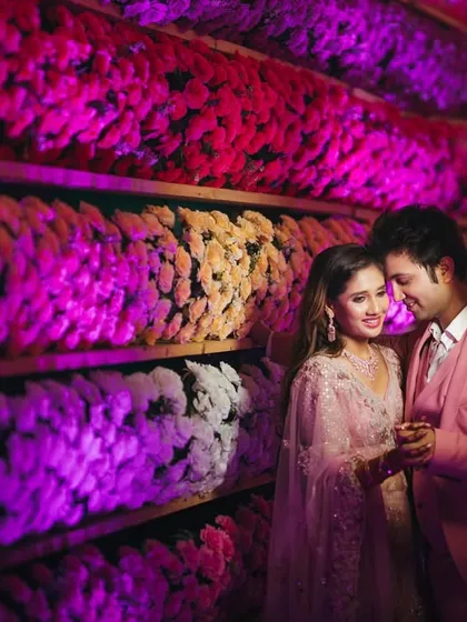 An intimate moment shared against a wall of colorful flowers. The dramatic lighting highlights the couple's connection, with the vibrant decor creating a rich and textured background for their portrait.