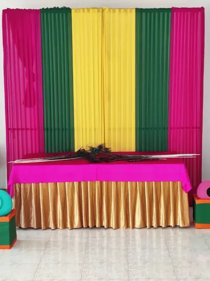 I create thematic decorations for specific festivals, like this pooja corner for a Janamashtmi celebration. The setup includes a colorful backdrop, a decorated table with a golden skirt for the idols, and traditional props like morpankh (peacock feathers) and matkis (clay pots).