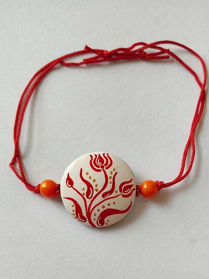 A beautiful terracotta rakhi with a delicate red floral painting on a white base, inspired by Bengali alpona. It's an elegant and artistic option.