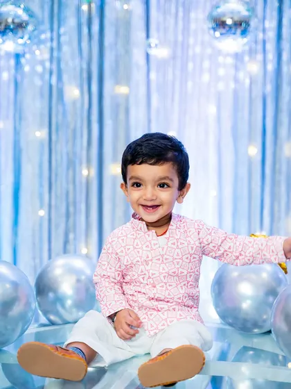 The birthday boy sits happily among silver balloons, dressed in a traditional pink and white kurta for his first birthday party.