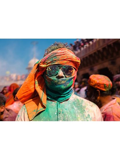 A man at the Holi festival, his face covered in green and orange powder, wearing sunglasses. This portrait captures the cool, modern attitude that coexists with ancient traditions.