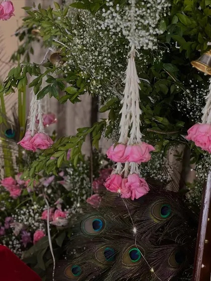 A close-up showing the beautiful details of our Janmashtami decor. Peacock feathers are tucked into the greenery behind hanging floral tassels, a symbolic nod to Lord Krishna.