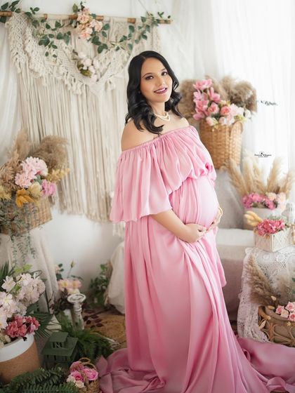 A beautiful solo portrait of a mother-to-be in a flowing pink off-shoulder gown. She looks radiant and serene in our flower-filled studio setup.