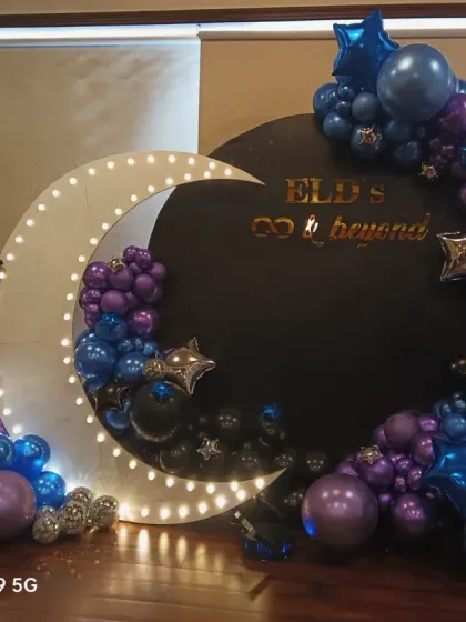 A celestial 'To the moon and beyond' theme. This decoration uses a black backdrop with a large light-up moon and a galaxy-colored balloon arrangement in purple, blue, and black.