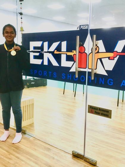 Our gold medal winner, Leno Caroline, proudly posing with her trophy and medal in front of the Eklavya Sports Shooting Academy entrance.
