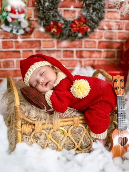 A newborn dressed as one of Santa's helpers, sleeping on a tiny bed with a ukulele, set against a cozy Christmas fireplace backdrop.