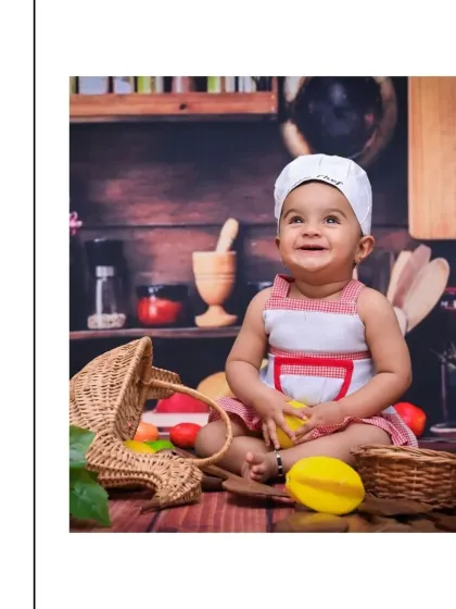 Looking up with a bright, happy smile, this little chef is surrounded by colorful toy vegetables and kitchen props. The rustic wooden background adds warmth to this adorable culinary-themed shoot.