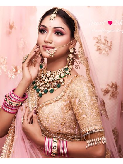 A dreamy bridal portrait with a soft pink background. The bride is wearing a heavy Polki and emerald choker, a delicate nath, and a simple maang tikka.