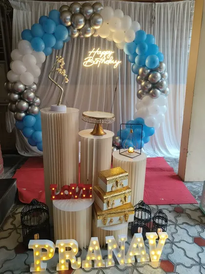 A classic blue, white, and silver balloon arch decoration for Pranav's birthday. The setup includes a neon sign, light-up letters, and multiple pedestals with props.