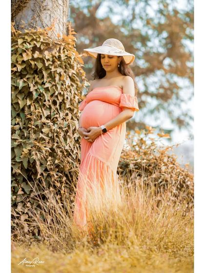 A full-length view of the mother-to-be in a beautiful off-shoulder gown, complemented by a stylish hat. The natural setting with ivy and tall grass adds texture and depth to the portrait.