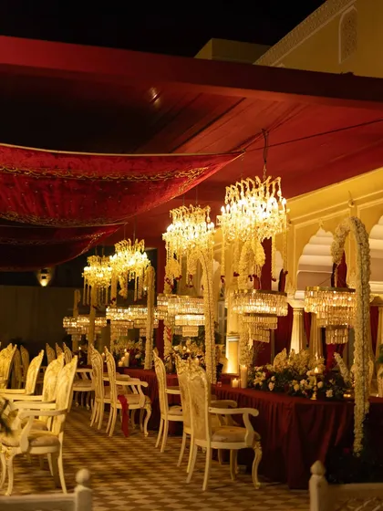 A long view of the royal dining setup, with tables draped in red velvet, adorned with chandeliers, and set in a checkered-floor courtyard.