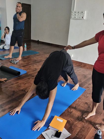 A student receives an adjustment in Downward-Facing Dog during a teacher training practicum, learning through direct experience.