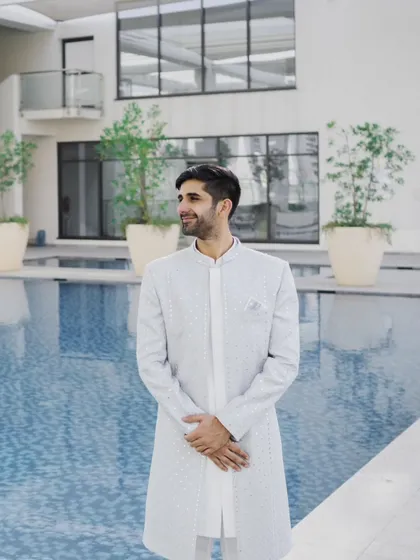 A portrait of the groom looking dapper in his light-colored sherwani, standing by the poolside of a modern hotel venue before his ring ceremony.
